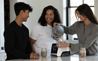 Three friends smile and chat in a kitchen as one of them pours a smoothie from a blender into a jar.