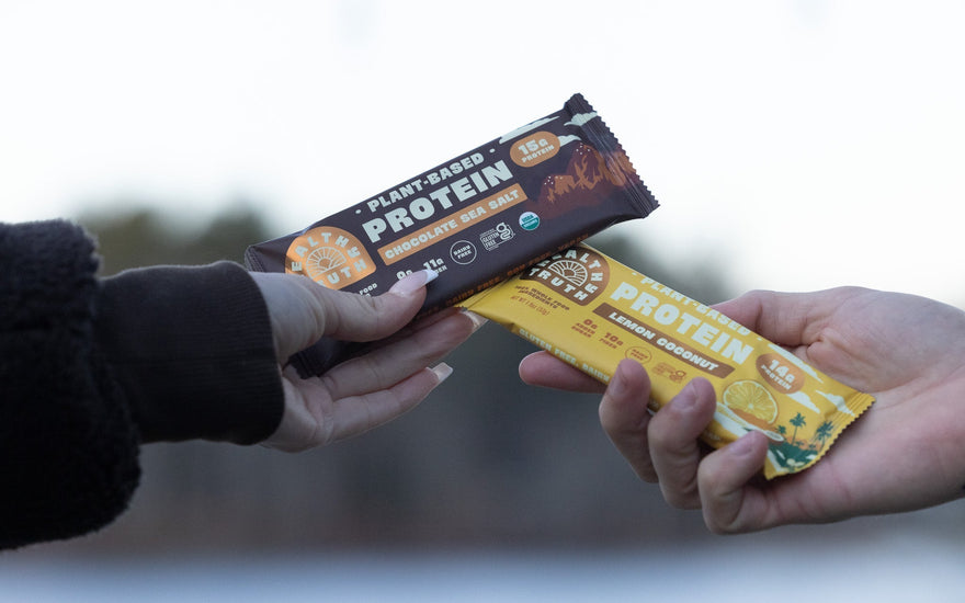 two hands each holding a protein bar in front of a blurred lake background