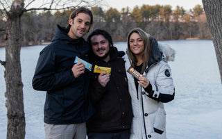 three people, two men and one woman in winter clothes each holding a protein bar in front of a lake