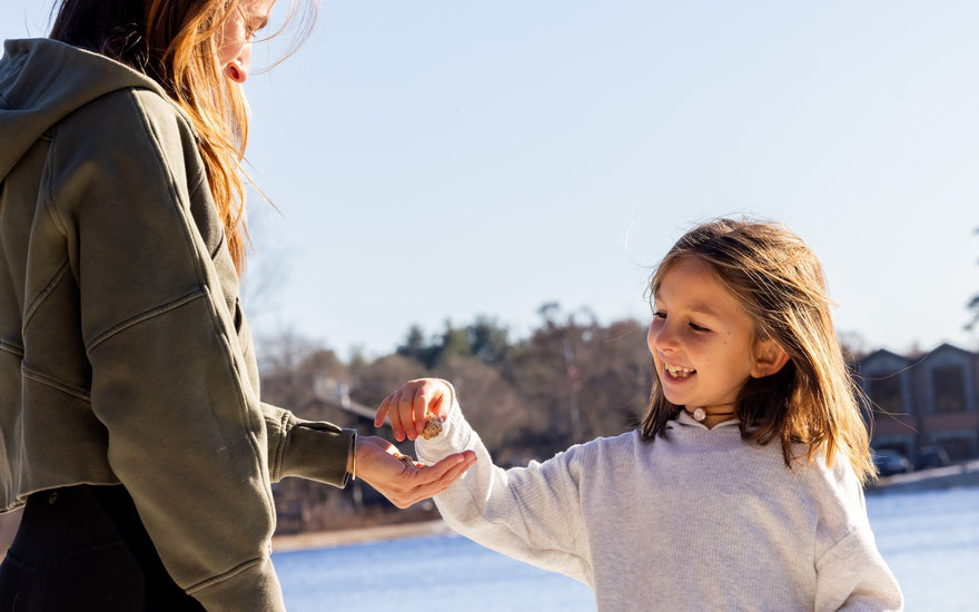 A smiling young girl takes a snack from a woman's hand on a sunny lakeside day.