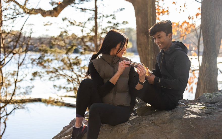 A man and woman sit on a rock outdoors by a lake, smiling and holding a protein bar together, surrounded by trees and sunlight.