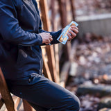 A person in athletic clothing leaning on a wooden bridge railing, outdoors in a natural setting, opening a blue-wrapped plant-based protein bar with visible icons and branding.