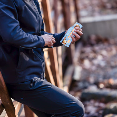 A person in athletic clothing leaning on a wooden bridge railing, outdoors in a natural setting, opening a blue-wrapped plant-based protein bar with visible icons and branding.