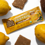 Flat lay of a Healthy Truth Lemon Coconut Plant-Based Protein bar surrounded by fresh lemons, a coconut, and pieces of the protein bar on a white background. Packaging highlights 14g protein, 11g fiber, no natural flavors, and USDA Organic certification.