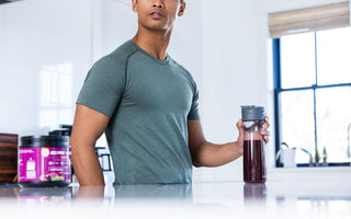 A man in a green athletic shirt holds a protein shake in a modern, bright kitchen.