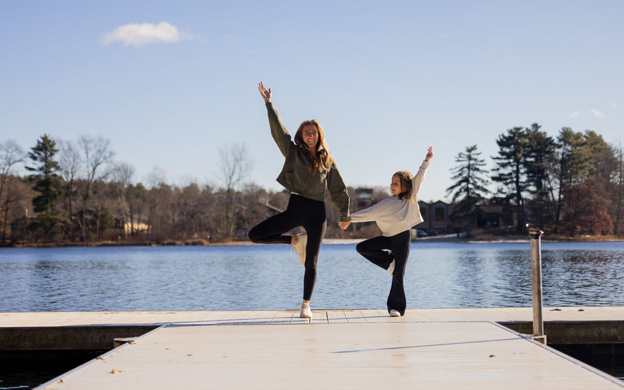 Two people, an adult and a child, pose in a yoga tree pose on a dock by a lake, smiling and holding hands on a clear day.