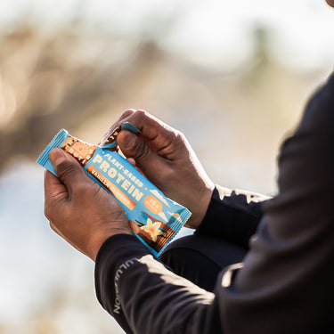 Close-up of a person opening a blue and brown protein bar labeled 'Plant-Based Protein – Vanilla Almond' with 15g of protein, sitting outdoors wearing a black athletic jacket.
