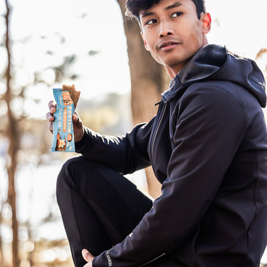 A man in athletic wear sitting on a rock outdoors, holding a partially unwrapped vanilla almond protein bar and looking off to the side with trees in the background.