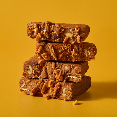 A stack of two halved chocolate sea salt protein bars broken in half to reveal a dense, crumbly texture with visible nuts and seeds, set against a vibrant yellow background.