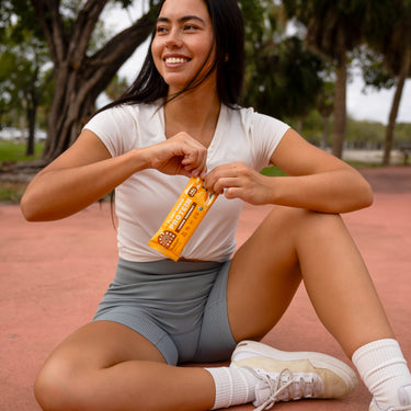 Young woman in athletic wear sitting on an outdoor court, smiling while opening a Lemon Coconut protein bar.