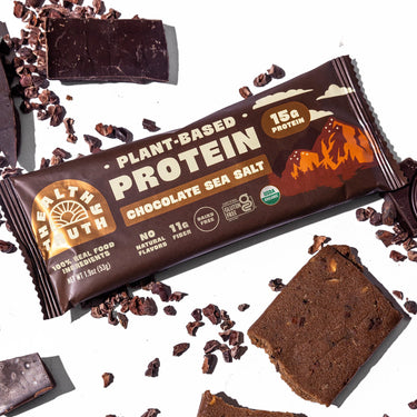 A Healthy Truth plant-based protein bar in Chocolate Sea Salt flavor on a white background, surrounded by chunks of chocolate and cocoa nibs, with the wrapper highlighting key features like 15g protein, 11g fiber, organic certification, and being gluten- and dairy-free.