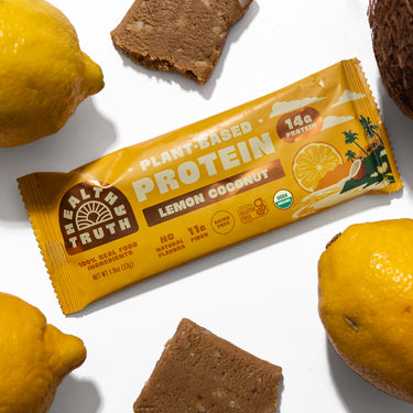 Flat lay of a Healthy Truth Lemon Coconut Plant-Based Protein bar surrounded by fresh lemons, a coconut, and pieces of the protein bar on a white background. Packaging highlights 14g protein, 11g fiber, no natural flavors, and USDA Organic certification.