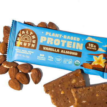 Flat lay of a Healthy Truth Vanilla Almond Plant-Based Protein bar surrounded by whole almonds and pieces of the protein bar on a white background. Packaging highlights 15g protein, 11g fiber, no natural flavors, and USDA Organic certification.
